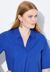 Blusa azzurra a maniche corte con scollo a V e un dettaglio pieghettato nella parte anteriore. Il modello indossa orecchini a goccia dorati. La texture appare liscia.