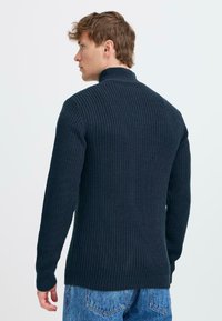 Navy blue knitted pullover sweater with a high collar, textured ribbing, and fitted sleeves, worn with faded blue denim jeans.
