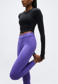 Γυναίκα που τεντώνει τον τετρακέφαλο, φοράει μαύρο crop top με μακριά μανίκια και μωβ κολάν New Balance, μπροστά σε λευκό φόντο.