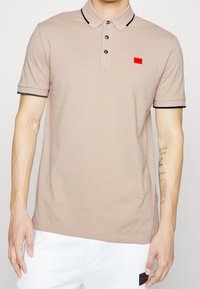 Polo shirt beige avec col et bords noirs, patte de boutonnage à trois boutons, et un petit logo rouge sur la poitrine. Matière en coton, texture lisse.