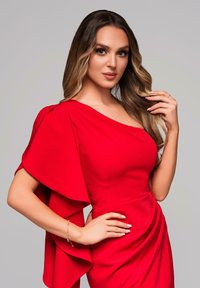 Robe du soir rouge avec un décolleté asymétrique, détail froncé sur le côté et manches surdimensionnées. Accompagnée d'un bracelet délicat.