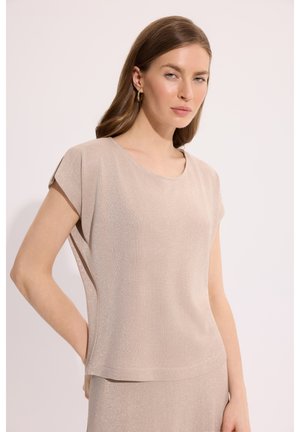 Kobieta o prostych brązowych włosach, ubrana w jasnobeżowy top z krótkim rękawem i pasującą spódnicę, stojąca na jednolitym tle.