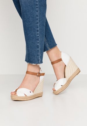 Pies con sandalias blancas de cuña con correas marrones en el tobillo y jeans azules recortados sobre un fondo claro y liso.