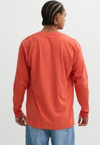 Camisa de manga larga en algodón color óxido, con un corte relajado, cuello redondo y mangas inseridas. Llevada con jeans de mezclilla en azul claro.