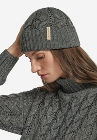 Szary dziergany beanie z teksturowanym wzorem warkocza, ściągaczem i beżową metką. Model nosi dopasowany szary sweter o wysokim kołnierzu z podobną teksturą.