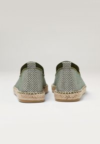 Espadrilles verdes a cuadros con parte superior de tejido entrelazado, suela de goma y detalles de cuerda de yute alrededor de la base. La vista trasera muestra una construcción resistente.