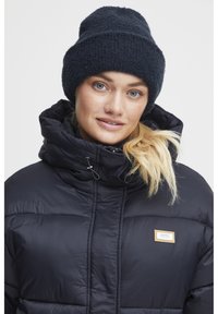 Giacca a vento blu navy con collo alto, design trapuntato e chiusura con zip. Abbinata a un berretto in maglia blu navy. Etichetta sulla giacca visibile.