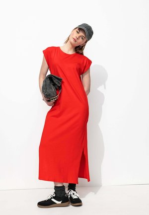 Robe maxi en coton rouge avec des manches courtes, fente latérale, associée à des baskets noir et blanc, des chaussettes sombres et une casquette grise à motifs.