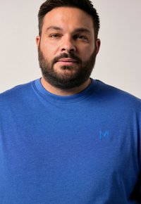 Muž s krátkými tmavými vlasy a vousy, oblečený v modrém tričku s kulatým výstřihem a malým logem "M" na levé straně hrudi, na jednobarevném pozadí.