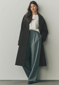 Svart trenchcoat över en vit skjorta, grå veckade byxor och svarta skor. Slät tygkvalitet, skräddarsydd passform och klassiska designelement.
