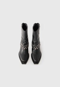 Botas de cuero negras hasta la mitad de la pantorrilla con punta cuadrada, adornadas con tachuelas de plata agrupadas alrededor del tobillo y el eje superior.