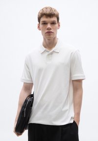 Fehér galléros póló klasszikus nyakkal és finom márkajelzéssel. A modell egy fekete táskát tart, amely cipzárral és strukturált kialakítással rendelkezik.