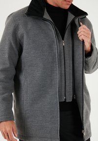 Abrigo de lana gris con cuello de piel sintética negra, cierre frontal con cremallera y dos bolsillos laterales. Cuenta con una chaqueta interior superpuesta. Textura suave.
