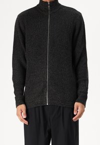 Dark grey zip-up knit sweater with a high collar, textured ribbed pattern, and long sleeves, worn over black trousers.