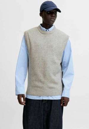 Gilet sans manches gris clair en maille douce, porté sur une chemise à rayures bleues et blanches avec des boutons, associé à un pantalon en denim foncé.