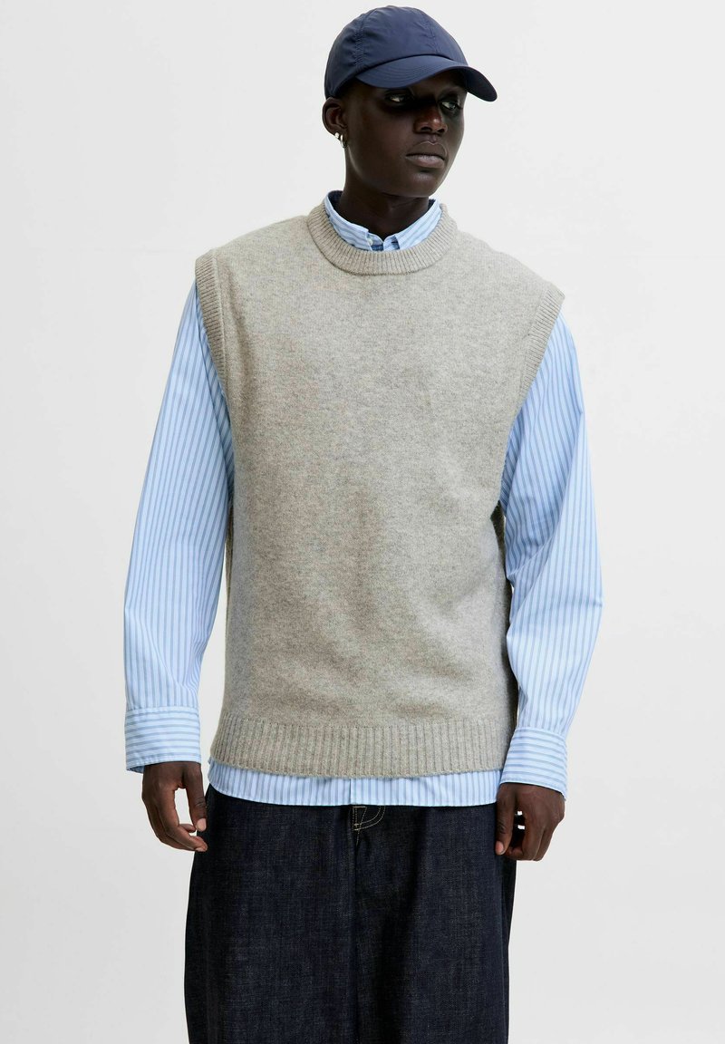 Gilet sans manches gris clair en maille douce, porté sur une chemise à rayures bleues et blanches avec des boutons, associé à un pantalon en denim foncé.