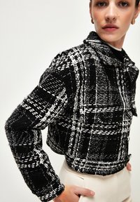 Chaqueta recortada de tweed en blanco y negro con cuello, con un patrón de cuadros, textura deshilachada y dos bolsillos frontales.