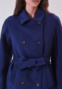 Cappotto doppiopetto blu navy in tessuto testurizzato, dotato di grandi bottoni, colletto e una cintura abbinata in vita con tasche laterali.