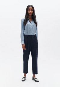 Himmelblaue Satinbluse mit Knopfschluss, kombiniert mit hochgeschnittenen marineblauen Hosen und schwarzen Loafers mit einem weißen Streifen.