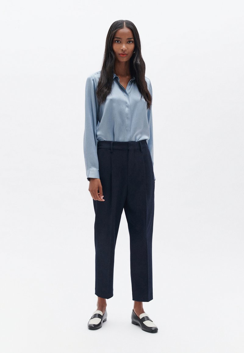 Himmelblaue Satinbluse mit Knopfschluss, kombiniert mit hochgeschnittenen marineblauen Hosen und schwarzen Loafers mit einem weißen Streifen.