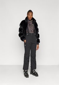 Casaco de pelo sintético preto com mangas listradas, por cima de uma blusa de estampado de leopardo castanho, combinado com calças de neve pretas e botas de esqui, a posar contra um fundo simples.