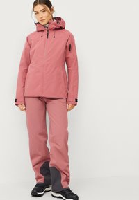 Giacca impermeabile rosa con cappuccio e cerniere, abbinata a pantaloni rosa coordinati con dettagli grigio scuro alle caviglie. Calzature nere.