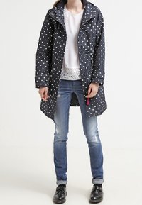 Impermeabile a pois navy con cappuccio, maniche lunghe e zip frontale. Indossato sopra una maglietta bianca e jeans blu, con scarpe nere.