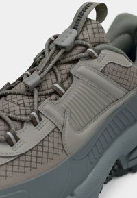 Beige sporta apavi ar teksturētu augšdaļu, elastīgām auklām un cietu gumijas zoli. Iezīmējas ar pelēku akcentu un izteiktu logo detaļu.