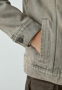 Gaiši pelēka džinsa jaka ar pogas aizdari uz aproces un sānu kabatu, teksturētu audumu un klasiskām dizaina detaļām.