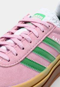 Rosa mockaskor med tre gröna ränder, vit tunga och beige gummisula. Har strukturerade snörningar och "GAZELLE"-logotyp.