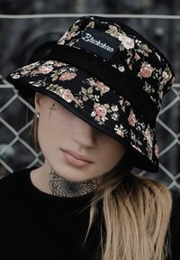 Cappello a secchiello nero con motivo floreale in rosa e verde su sfondo nero. Presenta una visiera ampia e un'etichetta in tessuto sulla parte anteriore.