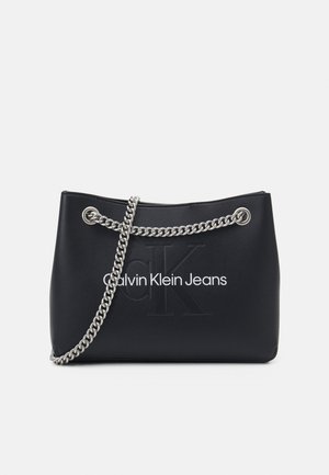 Czarna torebka Calvin Klein Jeans z srebrnym łańcuszkiem i wytłoczonym logo ck na gładkiej powierzchni na białym tle.