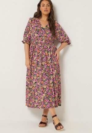 Kobieta stojąca, ubrana w kolorową sukienkę midi w różowo-pomarańczowo-żółty kwiatowy wzór oraz brązowe sandały z paskiem na kostkę, na tle jednolitej ściany.