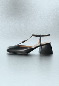 Μαύρο δερμάτινο παπούτσι με χαμηλό χοντρό τακούνι, λουράκι σε σχήμα Τ, στρογγυλεμένη τετράγωνη μύτη και κλείσιμο με αγκράφα στον αστράγαλο, σε απλό γκρι φόντο.