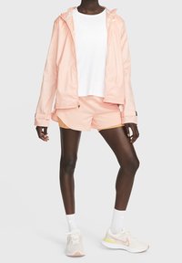 Lichtroze waterdichte jas met trekkoordcapuchon, over een wit T-shirt en bijpassende roze shorts. Witte sneakers met gele accenten.