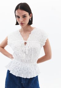 Blusa bianca texture con scatole, maniche corte, scollatura a chiave e vita aderente. Abbinata a pantaloni corti in denim blu.