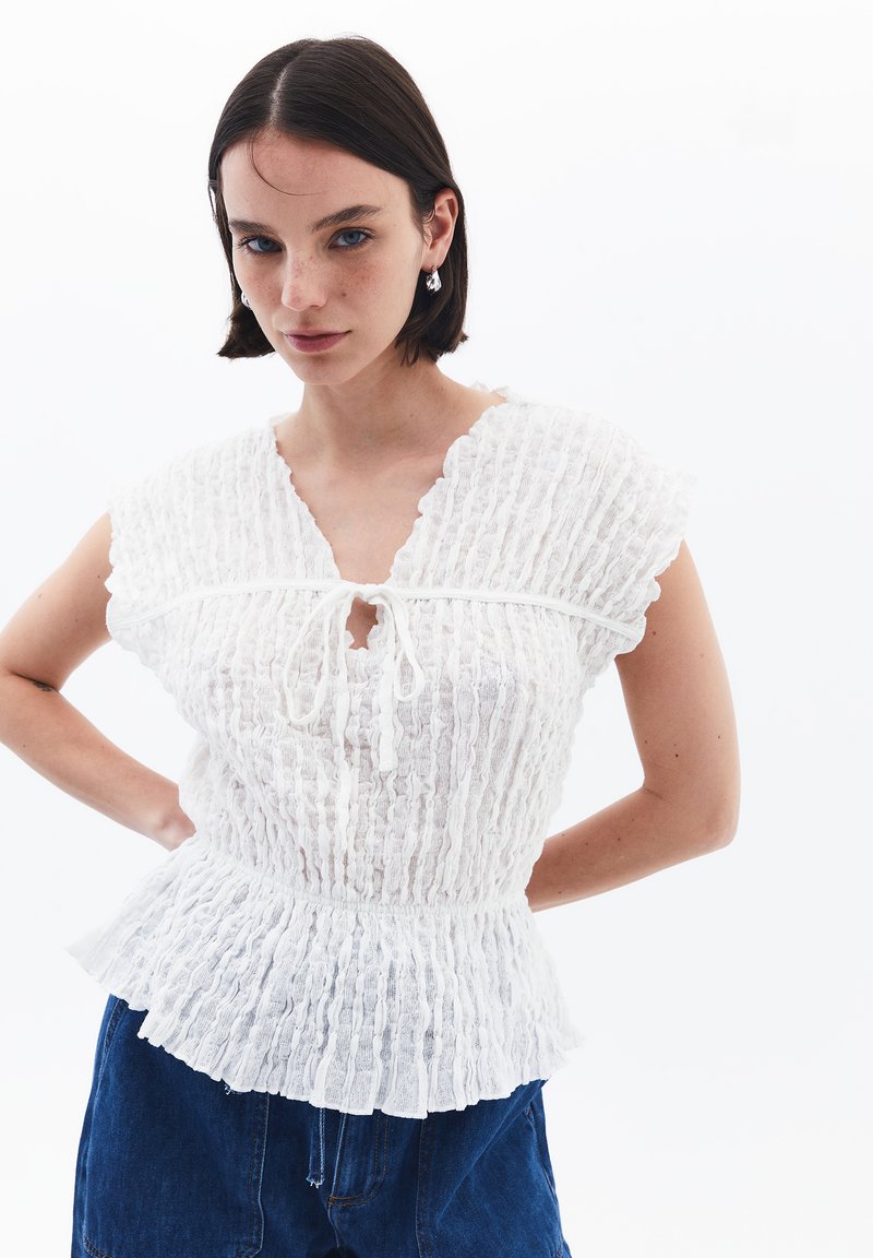 Blusa bianca texture con scatole, maniche corte, scollatura a chiave e vita aderente. Abbinata a pantaloni corti in denim blu.