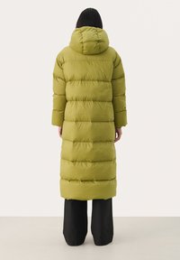 Olijfgroene pufferjas met een capuchon, voorzien van horizontale naden en een lange, cilindervormige pasvorm, gedragen over zwarte broek.