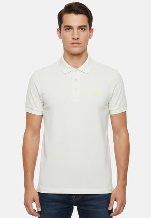 Giovane uomo che indossa una polo bianca a maniche corte con tre bottoni e un logo giallo-verde tenue sul petto e sulla manica.