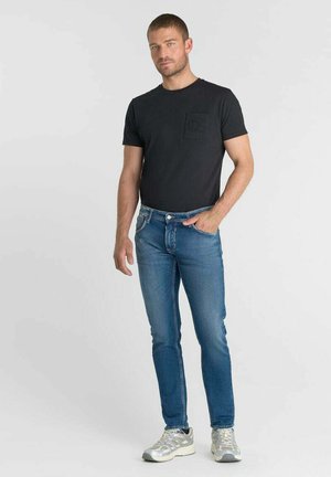 Uomo in piedi che indossa una t-shirt nera a maniche corte, jeans blu e sneakers bianche su uno sfondo grigio chiaro uniforme.