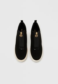 Scarpe slip-on nere in maglia con un dettaglio di cerniera dorata e un ciondolo logo. Presentano una tomaia testurizzata, una suola bianca a contrasto e un tirante sul tallone.