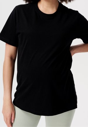 Person iført en ensfarvet sort kortærmet t-shirt og lyse ribstrikkede bukser, med den ene hånd på hoften og den anden afslappet ned langs siden.