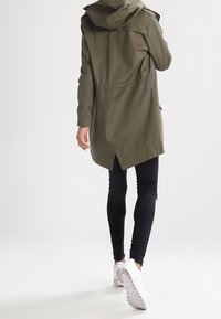 Parka vert olive avec capuche, taille cintrée et poches latérales ; associée à des leggings noirs et des baskets blanches.