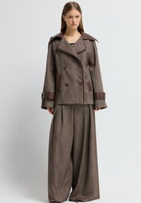 Giacca marrone a doppio petto con dettagli in pelle marrone e ampi polsini, abbinata a pantaloni larghi coordinati, entrambi con un motivo testurizzato.