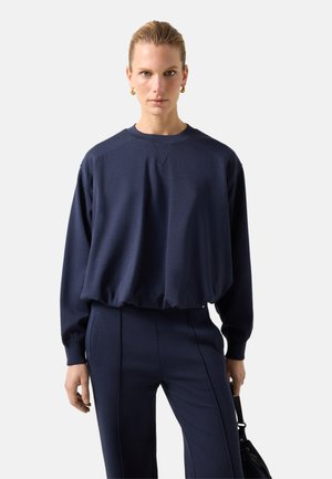 Kvinde iført en navyblå oversized sweatshirt og matchende bukser, der holder en sort håndtaske med store guld øreringe, neutralt baggrund.