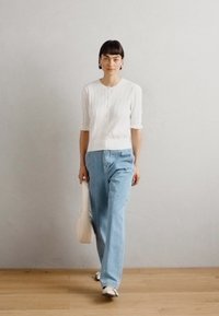 Cardigan bianco a coste con maniche corte, abbinato a jeans azzurri chiari. La modella porta una borsa beige chiaro e indossa sneaker bianche.