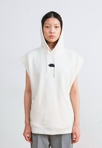 Beige ärmelloses Hoodie mit Kapuze und Kordelzug, mit einem kleinen schwarzen grafischen Detail auf der Brust. Weicher Stoff.