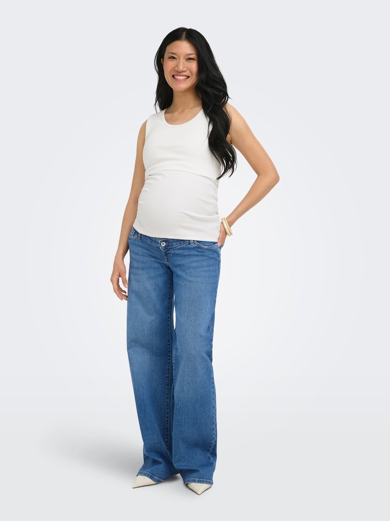 Femme enceinte souriante, portant un haut blanc sans manches, un jean bleu à jambes larges, des chaussures blanches et un bracelet, debout devant un fond uni.