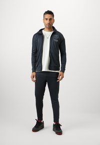 Mustad musta sportlik sättega jakk logoga, valge T-särk, mustad kitsenevad püksid ja mustad tossud punaste detailidega. Sujuv kangas, istuv disain.