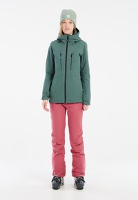 Grüne wasserdichte Jacke mit hohem Kragen, vorderen Reißverschlüssen und zwei Brusttaschen. Kombiniert mit rosa Skihosen und schwarzen Skistiefeln.